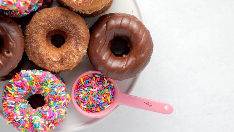 chocolate, plain and sprinkled doughnuts on plate tih pink spoon filled with multicolour sprinkles