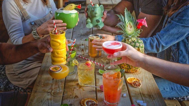 Group of people sat round an outside wooden bench doing a cheers with a variety of different coloured cocktails