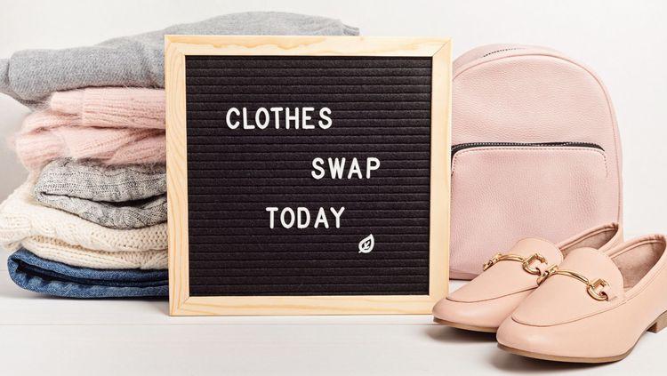 pile of multicoloured folders sweaters, a pink small backpack and shoes with a sign that says “Clothes Swap Today” in the centre in front of a white background