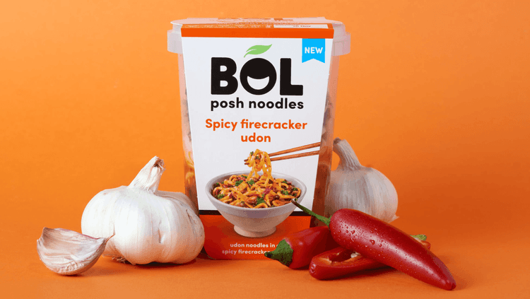 ready made noodles in a plastic container surrounded by garlic bulbs and chillies in front of an orange background 
