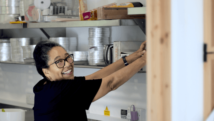 A smiling woman (Luzolo Ntima) reaching out to one of the shelves in her vegan bakery called The Heavenly Cake Co