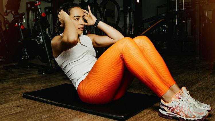 A woman in white top and orange with white and orange sports shoes sitting on a gym mat in exercising pose with dark background