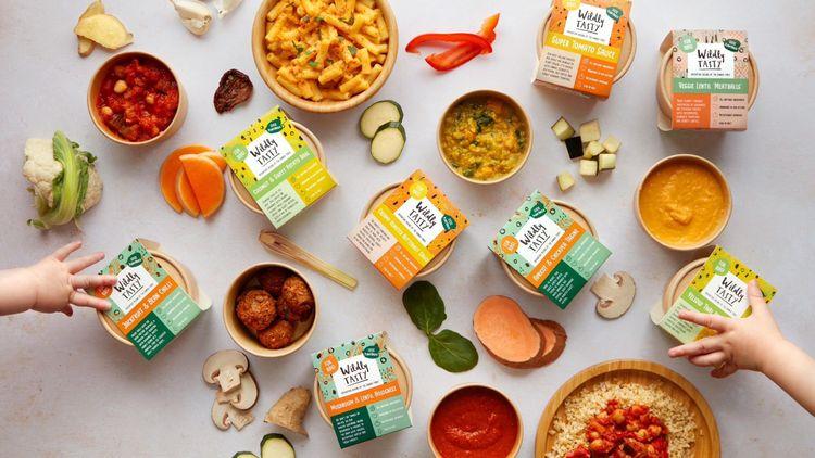 A variety of colourful plant-based foods by Wildly Tasty with their constituent fresh ingredients by the packs and two kids hands grabbing a pack of food