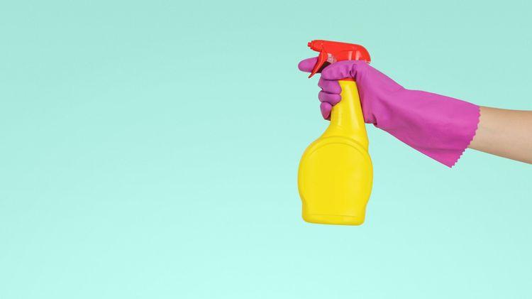 Bright yellow and red bottle of eco-friendly cleaning spray, held by a hand with a pink latex glove on spraying it on a blue background 