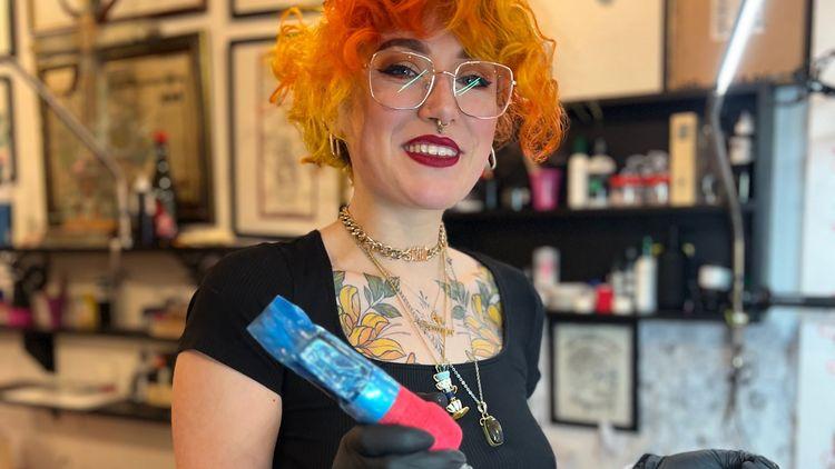 Vegan tattoo artist (Mogg) with brightly coloured hair smiling and holding a tattoo gun in a tattoo studio