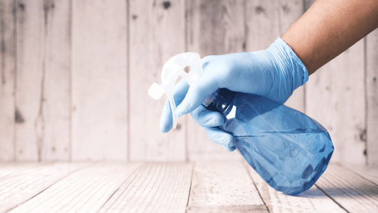 Person in a blue latex glove holding a transparent blue spray bottle over a wooden surface