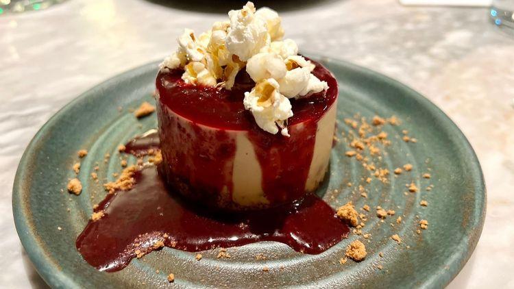 Vegan Caramel Cookie Cheesecake, drizzled with a raspberry coulis and topped with popcorn on a blue metallic plate in Mildreds Soho