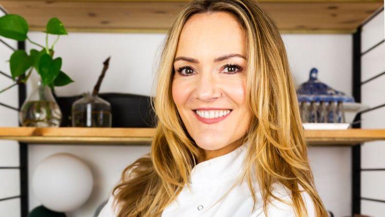Smiling vegan chef (Lisa Marley) in a chef’s white apron with blonde hair in front of a wooden shelf 