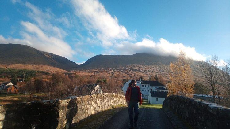 Man walking over a bridge (Bridge of Orchy) in the Scottish Highlands with a B&B in the background nestled beneath the mountains, trees and clouds