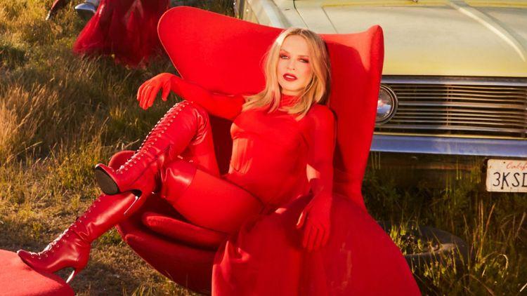Woman (Kylie Minogue) dressed fully in red on a red chair on long grass in front of an old car