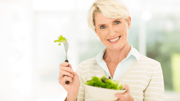 Smiling middle-aged woman holding a bowl full of salad leaves in one hand and a fork with a piece of salad in the other