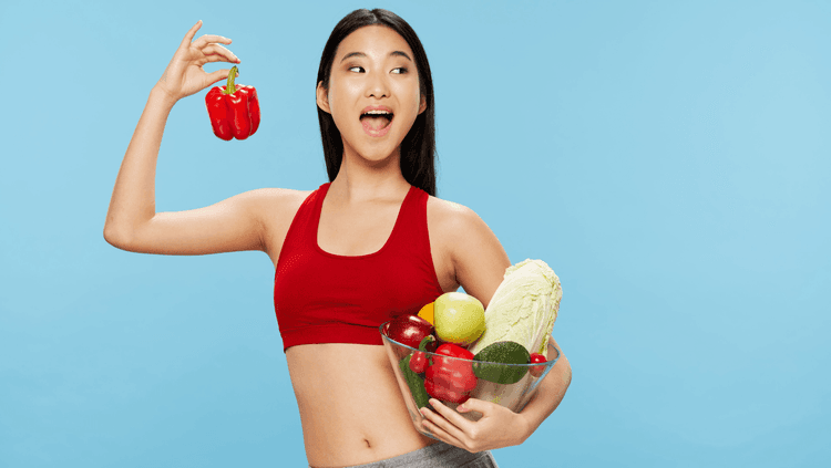 Smiling woman in a red top holding a red pepper in one hand and a glass bowl of vegetables in the other one on a blue background