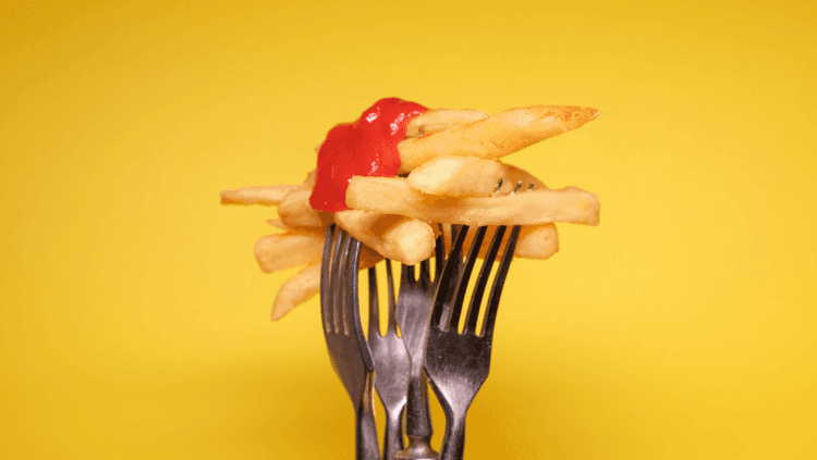 Chips covered with tomato ketchup being held up by forks on a yellow background.